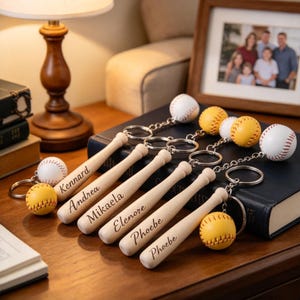 May include: Five wooden baseball bat keychains with metal rings, each featuring a small baseball or softball attached. The bats are engraved with the names Kennard, Andrea, Mikaela, Elenore, and Phoebe in a cursive script font.