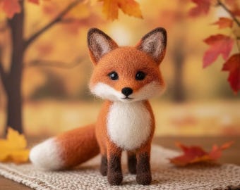 Puede incluir: Una figura de zorro naranja hecha a mano con pecho y punta de la cola blancos, y patas y orejas marrones oscuros. El zorro está de pie sobre una estera tejida beige, con un fondo otoñal borroso de hojas y árboles.