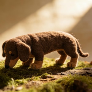 Könnte beinhalten: Handgefertigte Dackel-Figur aus Filz in Braun- und Beigetönen. Der Hund ist in einer Schnüffelpose dargestellt, mit detaillierten Merkmalen und einer strukturierten Optik. Die Figur steht auf grünem Moos. Ca. 20 cm lang.