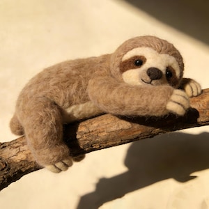 May include: A handcrafted sloth figure made from tan and brown wool, positioned on a wooden branch. The sloth features a light brown face with dark brown markings around the eyes and a small, dark brown nose. The sloth is in a relaxed, lying down pose.