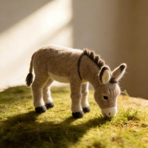 May include: A handcrafted felt donkey figurine in shades of tan and brown, with a dark mane and tail. The donkey is depicted grazing on green grass, with black hooves and white accents on its ears and muzzle. The figurine is a charming decorative item.