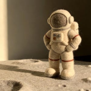 Puede incluir: Figura de astronauta hecha a mano en fieltro, con un traje espacial crema con detalles rojos y grises. El astronauta está de pie sobre una superficie gris texturizada, posiblemente representando la luna. La figura tiene un casco y una mochila detallados.
