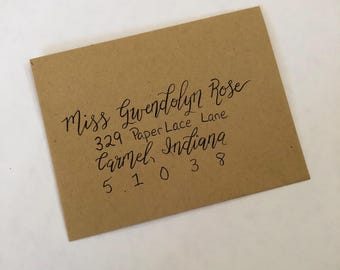 Puede incluir: Un sobre rectangular de color tostado con texto escrito a mano en caligrafía negra que dice: Miss Gwendolyn Rose, 329 Paper Lace Lane, Carmel, Indiana, 51038. El sobre tiene un acabado mate y liso, colocado en ángulo.