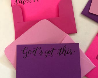 Puede incluir: Un sobre rosa brillante que contiene una tarjeta con el texto escrito a mano en negro trust in His timing, y un sobre rosa claro con una tarjeta morada con el texto escrito a mano en negro God's got this, con sobres adicionales.