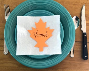 Puede incluir: Un juego de platos turquesa con una servilleta cuadrada blanca coronada por una tarjeta de sitio naranja con forma de hoja de arce que muestra el nombre Hannah en cursiva negra, flanqueado por un tenedor y un cuchillo de sierra.