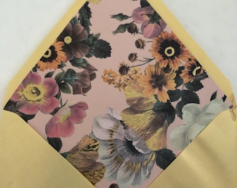Puede incluir: Un sobre abierto con un exterior dorado metálico y un forro interior rosa claro con un patrón floral detallado. El estampado muestra varias flores en tonos naranja, amarillo, rosa y blanco, acentuadas con detalles de lámina dorada.
