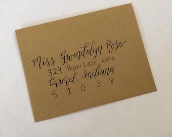 Puede incluir: Un sobre rectangular de color tostado con texto escrito a mano en caligrafía negra que dice: Miss Gwendolyn Rose, 329 Paper Lace Lane, Carmel, Indiana, 51038. El sobre tiene un acabado mate y liso, colocado en ángulo.