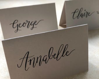 Puede incluir: Tres tarjetas de sitio plegadas de color beige claro hechas de cartulina. Cada tarjeta presenta un nombre escrito con tinta negra en letra cursiva. Los nombres visibles en las tarjetas son Annabelle, George y Claire, en el frente.