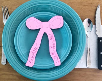 Puede incluir: Un recorte de papel rosa claro en forma de lazo con el texto "Caroline" y "a little bow-tiful" escrito en cursiva, colocado sobre dos platos turquesa, acompañado de un tenedor, una cuchara y un cuchillo de sierra de plata.