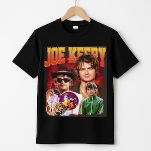 May include: Black t-shirt featuring a retro-style graphic of Joe Keery with the text "JOE KEERY" in orange and yellow. The design includes multiple images of the actor in various poses. The shirt is hanging on a wooden hanger.