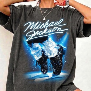 May include: Dark gray t-shirt featuring a graphic of Michael Jackson's iconic shoes and the artist's name in white script. The image has a blue and white spotlight effect, creating a stage-like atmosphere. The shirt is a casual, oversized style.