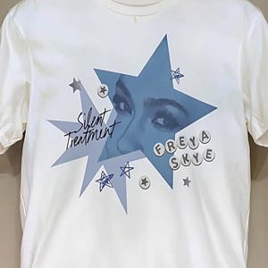 May include: Off-white t-shirt featuring a blue starburst design with a woman's eyes and the text "Silent Treatment." The name "FREYA SKYE" is spelled out in individual letter circles. Additional small star graphics are scattered around the design.