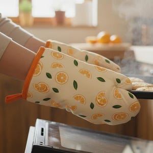 Puede incluir: Una persona con guantes de cocina con un diseño de rodajas de naranja saca una bandeja de galletas del horno. Los guantes son de color crema con detalles en naranja y verde. Sale vapor del horno.