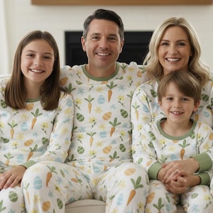 May include: A family of four wearing matching pyjamas. The pyjamas are white with a pattern of carrots, eggs, and flowers. The pyjamas have green trim around the neck and wrists. The family is sitting on a white sofa.