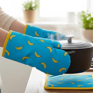 Puede incluir: Guantes de horno azules y un salvamanteles a juego con un estampado de plátanos amarillos. Los guantes se utilizan para levantar una olla negra con vapor. Los guantes y el salvamanteles tienen ribetes amarillos.