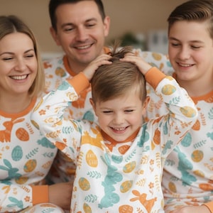 Puede incluir: Una familia de cuatro personas con pijamas blancos a juego con ribetes naranjas y un estampado de huevos y conejos de Pascua. La familia sonr&iacute;e y r&iacute;e, creando un ambiente alegre. Los pijamas parecen estar hechos de un material suave y c&oacute;modo.