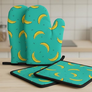 Puede incluir: Un par de guantes de horno turquesa y dos salvamanteles a juego. Los textiles de cocina presentan un patrón repetido de plátanos amarillos. Los guantes y salvamanteles tienen un ribete negro y están sobre una superficie de madera clara.