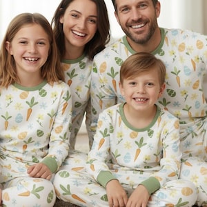 May include: A family of four wearing matching pyjamas. The pyjamas are white with a pattern of carrots, eggs, and flowers. The pyjamas have green trim on the collar and cuffs. The family is smiling at the camera.