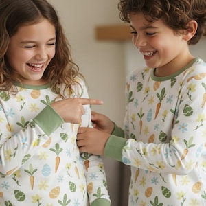 May include: Two children wearing matching white pyjamas with a colourful Easter-themed print. The long-sleeved pyjamas feature carrots, flowers, and Easter eggs. The pyjamas have green trim around the neck and cuffs. The children are smiling and pointing at each other.