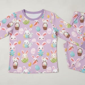 Puede incluir: Un conjunto de pijama lila con un estampado repetido de conejitos de Pascua, huevos y cestas. La parte superior de manga larga y los pantalones tienen pu&ntilde;os y cuello lila. El pijama est&aacute; dise&ntilde;ado para la comodidad y es adecuado para ni&ntilde;os.