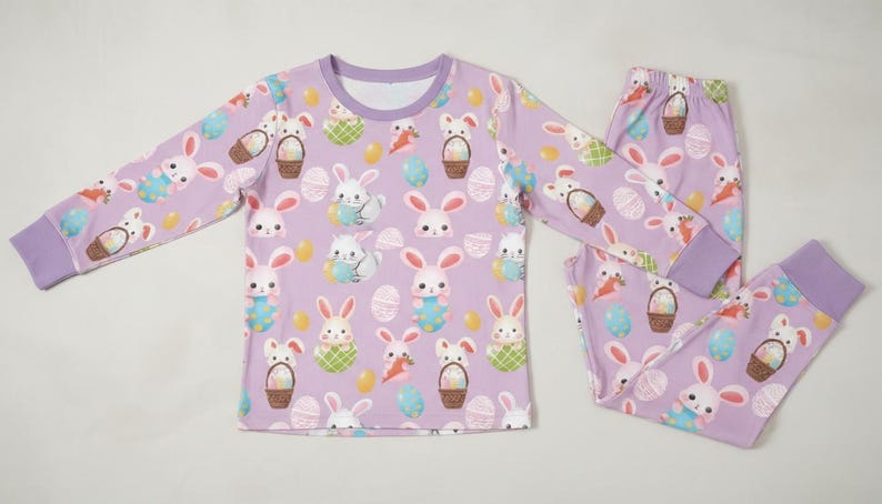 Puede incluir: Un conjunto de pijama lila con un estampado repetido de conejitos de Pascua, huevos y cestas. La parte superior de manga larga y los pantalones tienen pu&ntilde;os y cuello lila. El pijama est&aacute; dise&ntilde;ado para la comodidad y es adecuado para ni&ntilde;os.