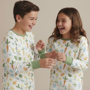May include: Two children wearing matching white pyjamas with a colourful Easter-themed print. The pyjamas feature carrots, flowers, and Easter eggs. The long-sleeved tops have green trim around the neck and cuffs.