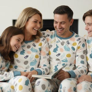 Puede incluir: Una familia de cuatro personas con pijamas a juego con un estampado de conejos y huevos. Los pijamas son blancos con detalles en azul, amarillo y rosa. Est&aacute;n sentados en un sof&aacute; gris, sonriendo y leyendo un libro.