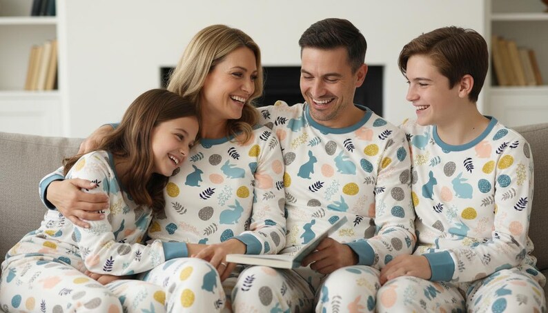 Puede incluir: Una familia de cuatro personas con pijamas a juego con un estampado de conejos y huevos. Los pijamas son blancos con detalles en azul, amarillo y rosa. Est&aacute;n sentados en un sof&aacute; gris, sonriendo y leyendo un libro.