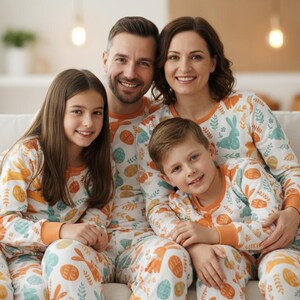 Puede incluir: Una familia de cuatro sonr&iacute;e sentada en un sof&aacute; blanco. Llevan pijamas a juego con un fondo blanco y un estampado de huevos naranjas, conejos azules y hojas verdes. Los pijamas tienen pu&ntilde;os naranjas.