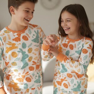 Puede incluir: Dos ni&ntilde;os con pijamas a juego con motivos de Pascua. Los pijamas son blancos con un estampado de conejos, huevos y follaje en naranja y azul. Las camisetas de manga larga tienen ribetes naranjas en el cuello y los pu&ntilde;os. Los ni&ntilde;os sonr&iacute;en y r&iacute;en.