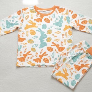 Puede incluir: Conjunto de pijama blanco con ribete naranja, con un estampado de conejitos naranjas y turquesas, huevos de Pascua y detalles florales. La parte superior de manga larga y los pantalones a juego est&aacute;n dise&ntilde;ados para la comodidad.