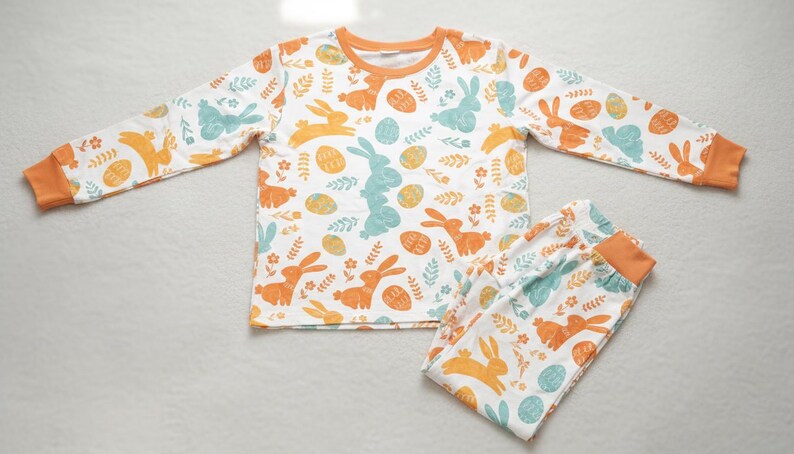 Puede incluir: Conjunto de pijama blanco con ribete naranja, con un estampado de conejitos naranjas y turquesas, huevos de Pascua y detalles florales. La parte superior de manga larga y los pantalones a juego est&aacute;n dise&ntilde;ados para la comodidad.