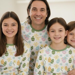 Pu&ograve; includere: Una famiglia di quattro persone indossa pigiami abbinati. I pigiami sono bianchi con un motivo di uova di Pasqua e fiori dai colori pastello. La famiglia sorride alla fotocamera.