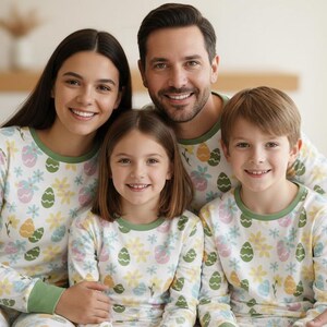 Pu&ograve; includere: Una famiglia di quattro persone indossa pigiami coordinati. I pigiami sono bianchi con un motivo di uova di Pasqua e fiori in colori pastello. I pigiami hanno finiture verdi intorno al collo e alle maniche.