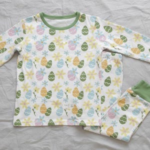 Pu&ograve; includere: Pigiama bianco con un motivo colorato di uova di Pasqua e fiori. La maglia a maniche lunghe e i pantaloni hanno bordi verdi. Il motivo include uova e fiori gialli, verdi, blu e rosa.