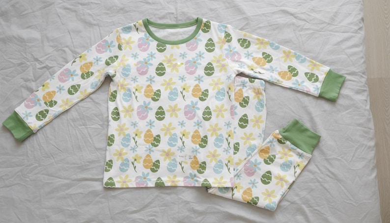 Pu&ograve; includere: Pigiama bianco con un motivo colorato di uova di Pasqua e fiori. La maglia a maniche lunghe e i pantaloni hanno bordi verdi. Il motivo include uova e fiori gialli, verdi, blu e rosa.