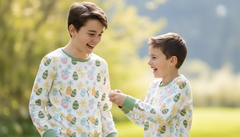 Pu&ograve; includere: Due persone indossano pigiami a maniche lunghe abbinati con un motivo di uova di Pasqua e fiori. I pigiami sono bianchi con finiture verdi e presentano uova e fiori dai colori pastello. Le persone sorridono e ridono all'aperto.