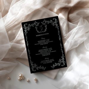 May include: A black rectangular wedding weekend events card featuring a white floral border and the initials A and J at the top. The text details Friday, Saturday, and Sunday events including times and attire requirements in a white serif font.