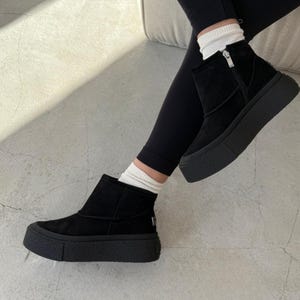 Puede incluir: Botines negros con suela de plataforma. Las botas tienen una cremallera lateral y están hechas de un material similar al ante. Las botas se usan con leggings negros y calcetines blancos.