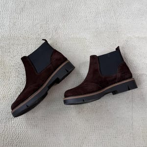 以下が含まれることがあります： 黒い伸縮性のあるサイドパネルと厚い黒いソールを備えた、茶色のスエードアンクルブーツのペア。 ブーツは丸いつま先と背面にプルタブが付いています。 ブーツは濃い茶色です。