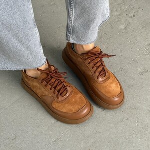 May include: A pair of brown suede and leather sneakers with matching laces. The shoes have a rounded toe and a thick, brown sole. The sneakers are worn with light blue jeans.