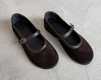 May include: A pair of dark brown Mary Jane shoes. The shoes feature a rounded toe, a strap with a buckle, and dark brown leather trim. They are made of suede and have a flat sole. The shoes are suitable for everyday wear.