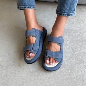May include: Blue suede sandals with adjustable straps. The sandals have a thick sole and a white footbed. The straps are secured with metal buckles. The sandals are worn with blue jeans.