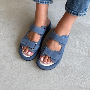 May include: Blue suede sandals with adjustable straps. The sandals have a thick sole and a white footbed. The straps are secured with silver-colored metal buckles. The sandals are worn with blue jeans.