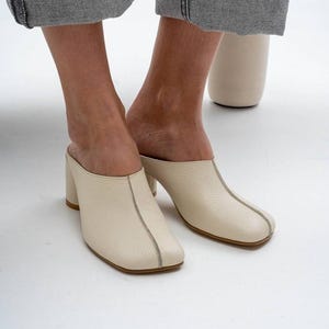 Puede incluir: Mules de cuero color crema con punta cuadrada y tacón de bloque. Los zapatos tienen un sutil detalle de costura y suela marrón claro. Los tacones miden aproximadamente 7,6 cm de alto.