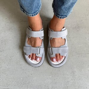May include: Gray suede sandals with two adjustable straps and a white footbed. The sandals have a minimalist design and are suitable for casual wear. The straps feature metal buckles for secure fastening.
