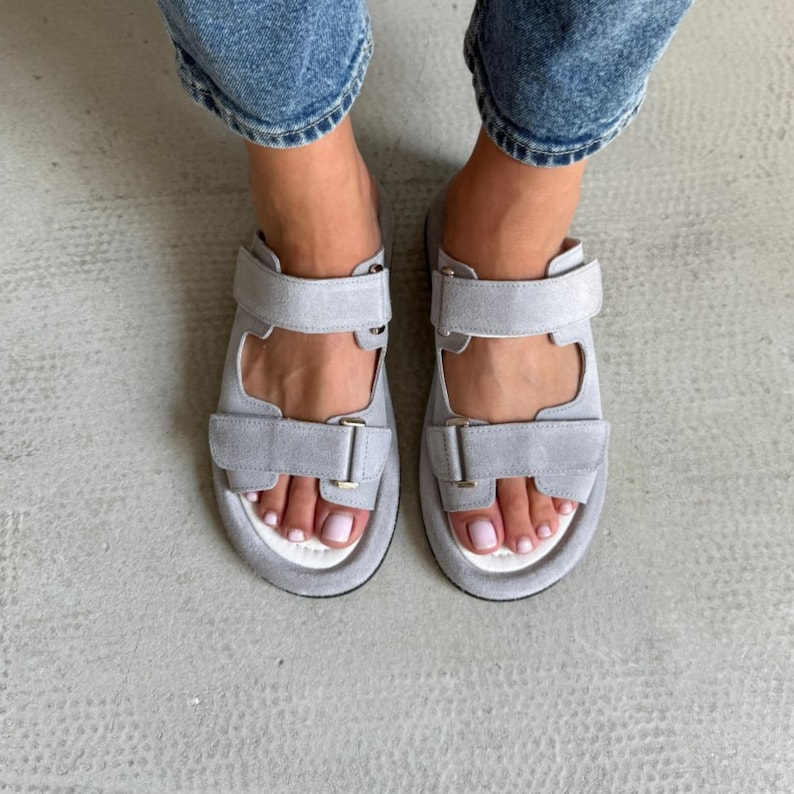May include: Gray suede sandals with two adjustable straps and a white footbed. The sandals have a minimalist design and are suitable for casual wear. The straps feature metal buckles for secure fastening.