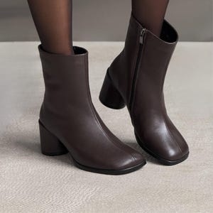 Puede incluir: Botines marrones oscuros con punta cuadrada y tacón grueso. Las botas tienen una cremallera lateral y están hechas de un material liso similar al cuero. Los tacones miden aproximadamente 7,6 cm de alto.