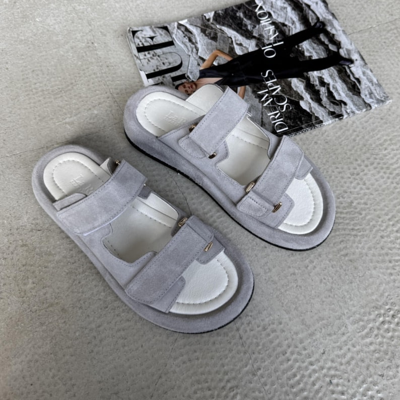 May include: A pair of light gray suede sandals with adjustable straps and white footbeds. The sandals have a thick sole and are displayed on a light gray surface. The sandals are a casual footwear option.
