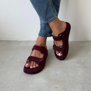 May include: Burgundy sandals with two adjustable straps and a thick sole. The sandals have a suede-like texture and feature silver-colored buckles. The footwear is worn with blue jeans.